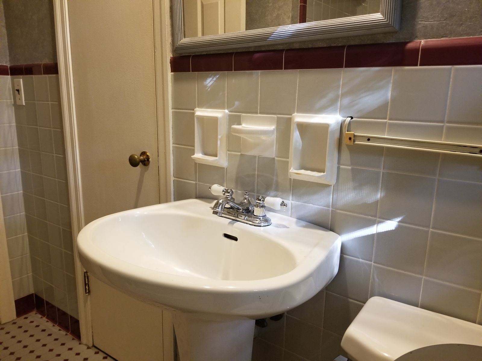 Original dated hall bath with a pedestal sink, gray and burgundy wall tile, ceramic soap dishes, and worn wallpaper