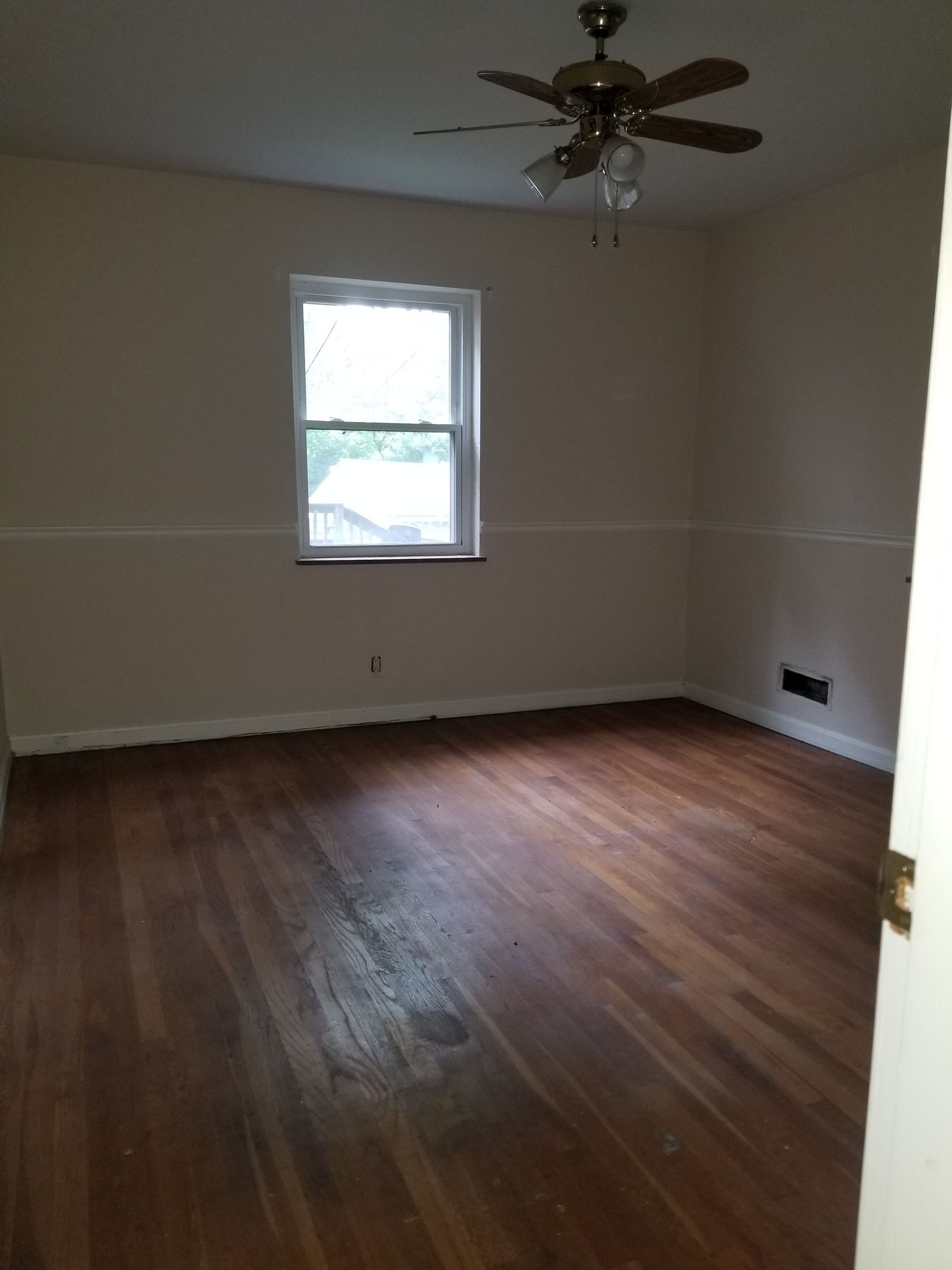 Tired main-floor bedroom with scuffed hardwood floors, patched walls, chair rail molding, and a dated brass ceiling fan