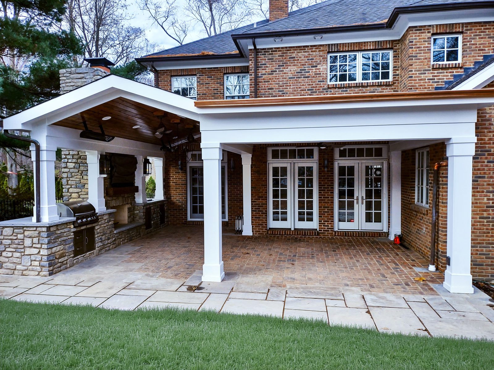 Gable-roofed covered porch with white columns, stone bases, and brick paver floor attached to a brick home