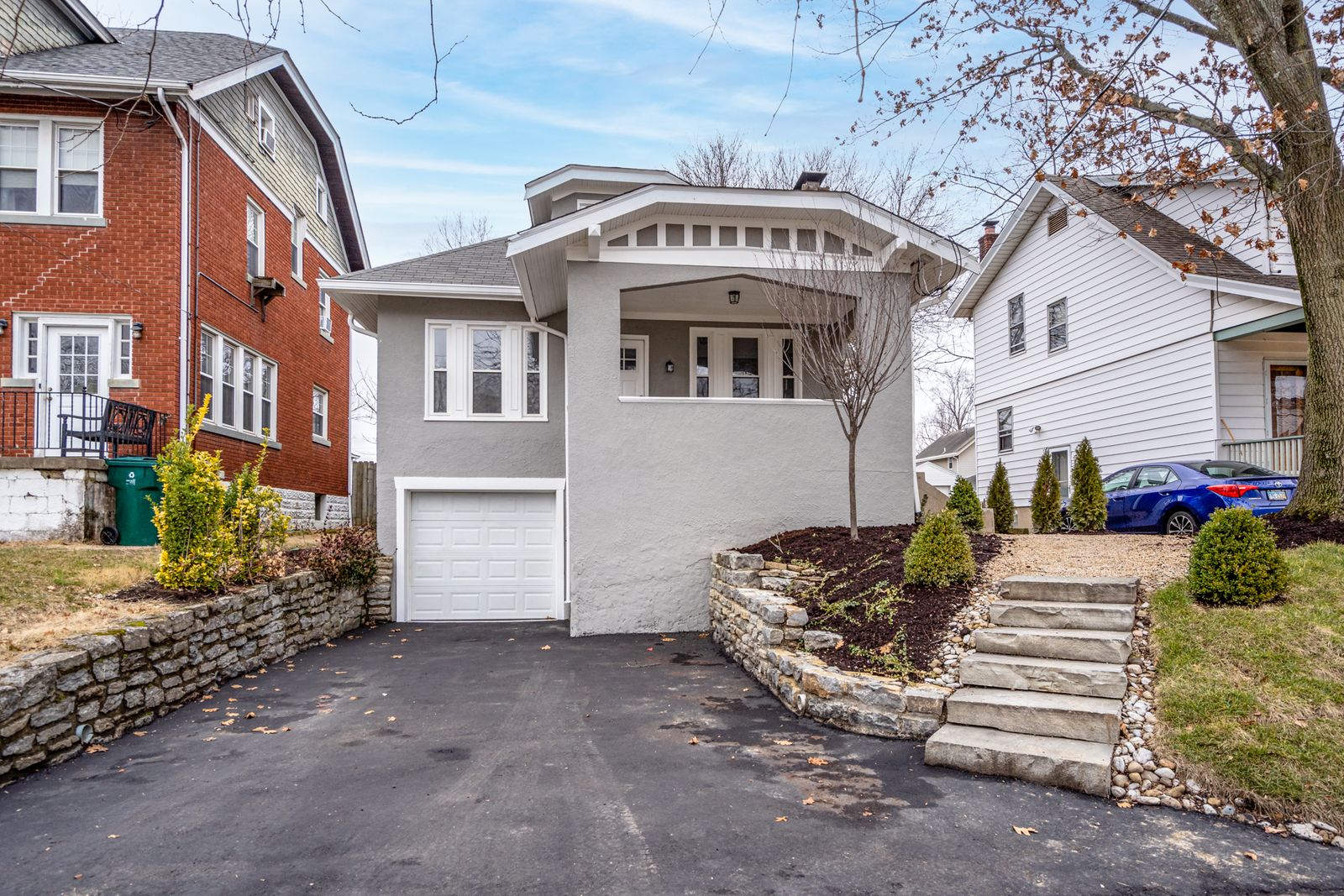 Front of a renovated gray stucco bungalow with white trim, covered entry porch, stone retaining walls, and a tuck-under garage