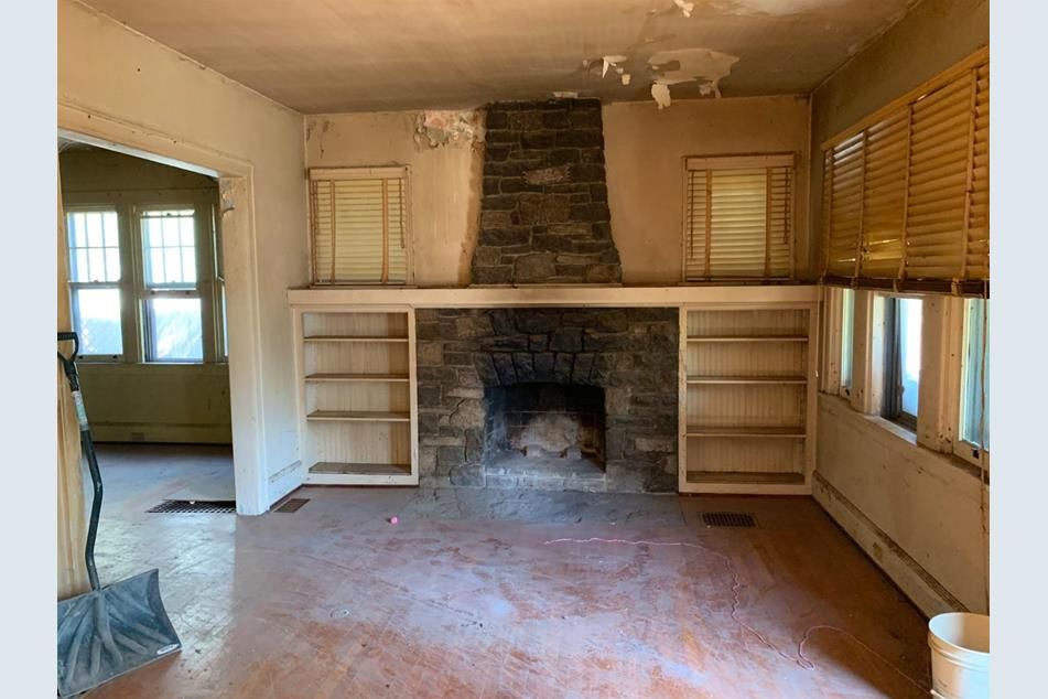 Living room before renovation, with a soot-stained stone fireplace, damaged plaster, a peeling ceiling, and worn floors